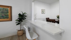 Un interior moderno con una pared blanca, un cuadro enmarcado, una planta en maceta, una silla blanca y una zona de comedor con una mesa y sillas.