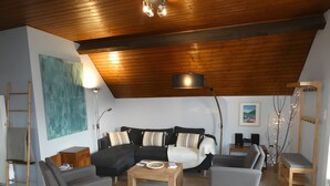 Un salon moderne avec un plafond en bois, un canapé d’angle, deux fauteuils, une table basse et une lampe.