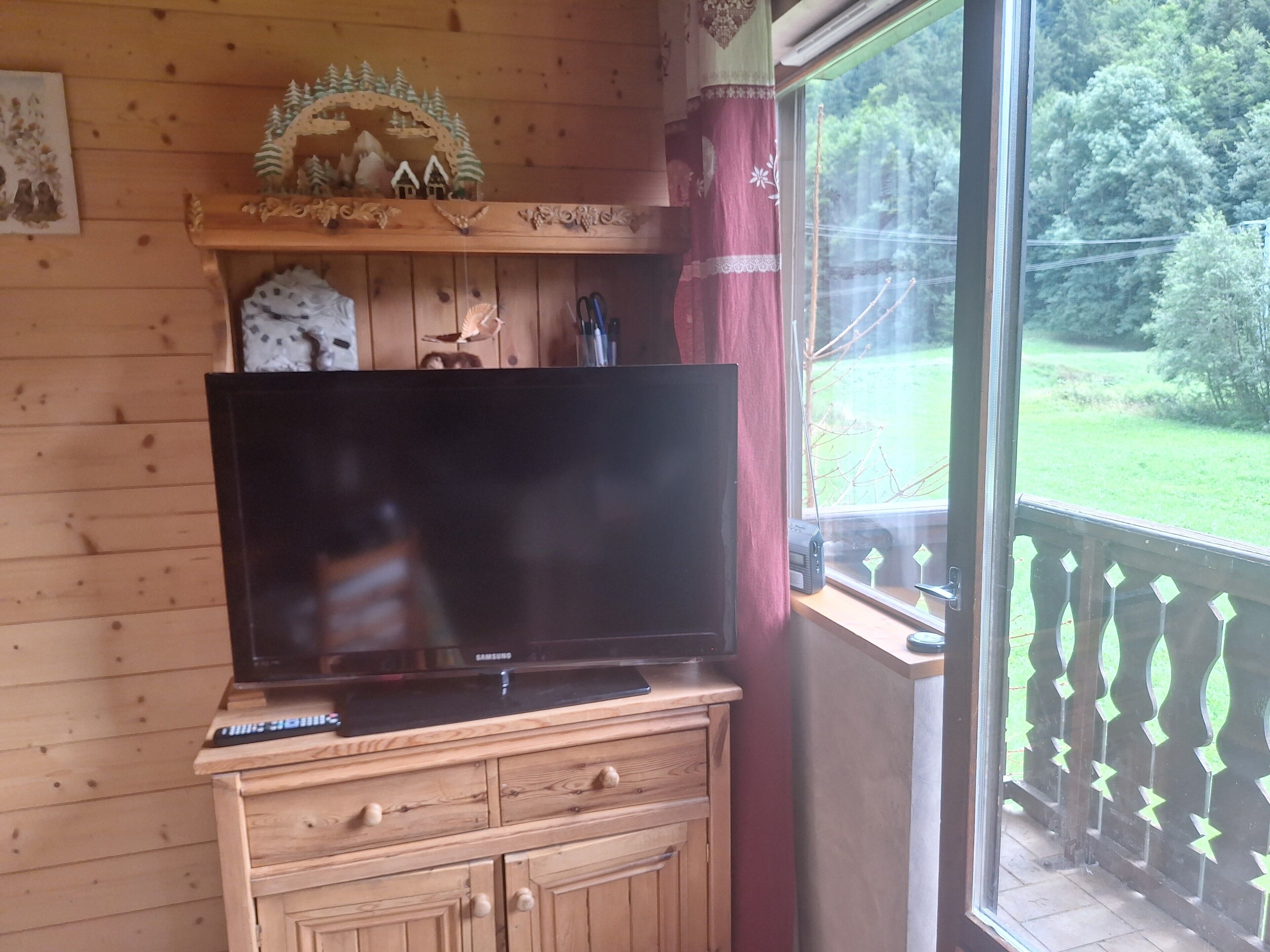 A wooden cabin interior with a television on a wooden cabinet, a decorative shelf above, and a view of a green landscape through a window.