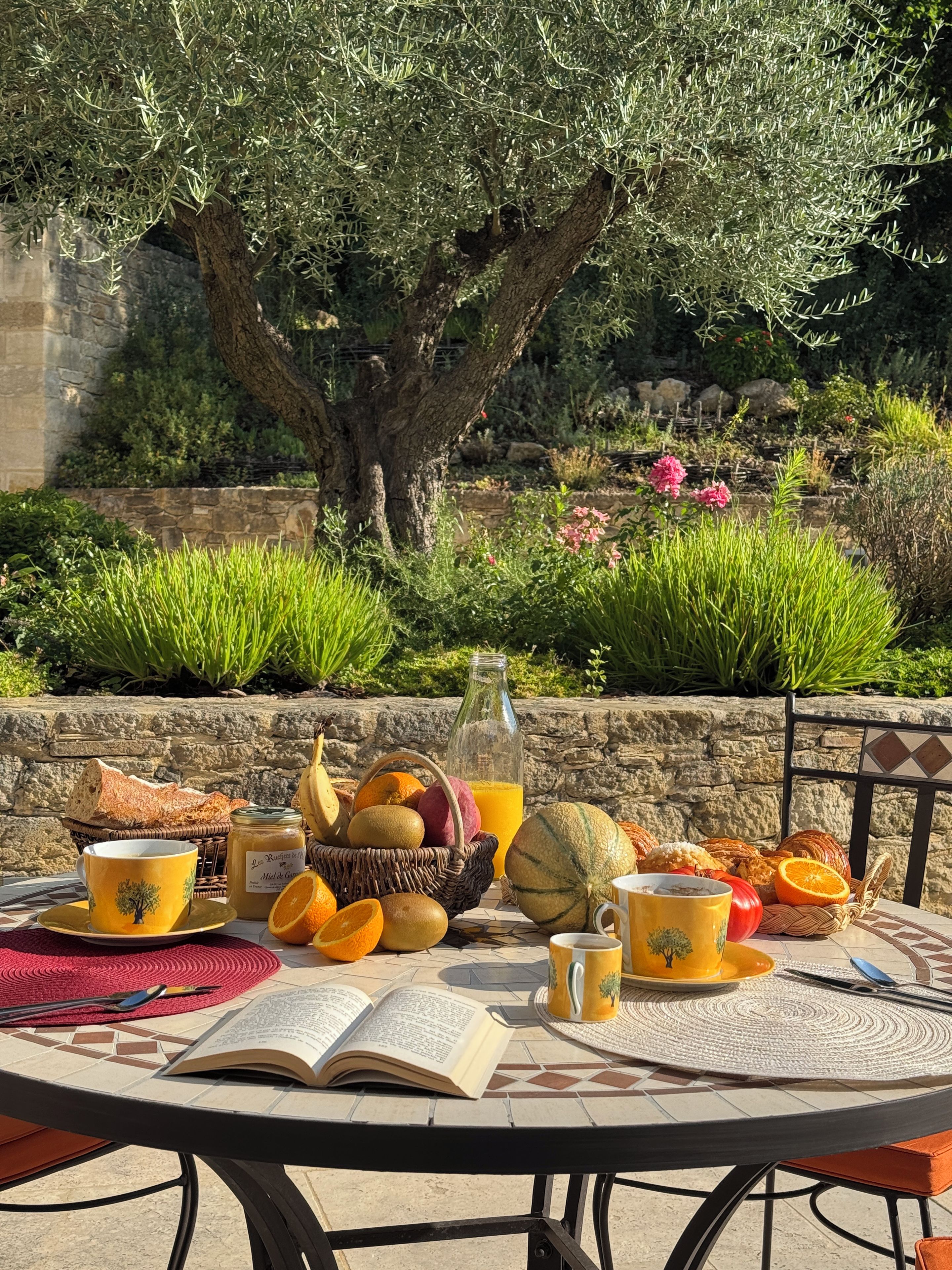 An outdoor dining table with a book, a basket of fruits, a bottle of juice, and a cup.