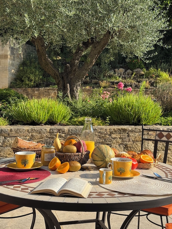 Une table à manger en plein air, avec un livre, un panier de fruits, une bouteille de jus et une tasse.