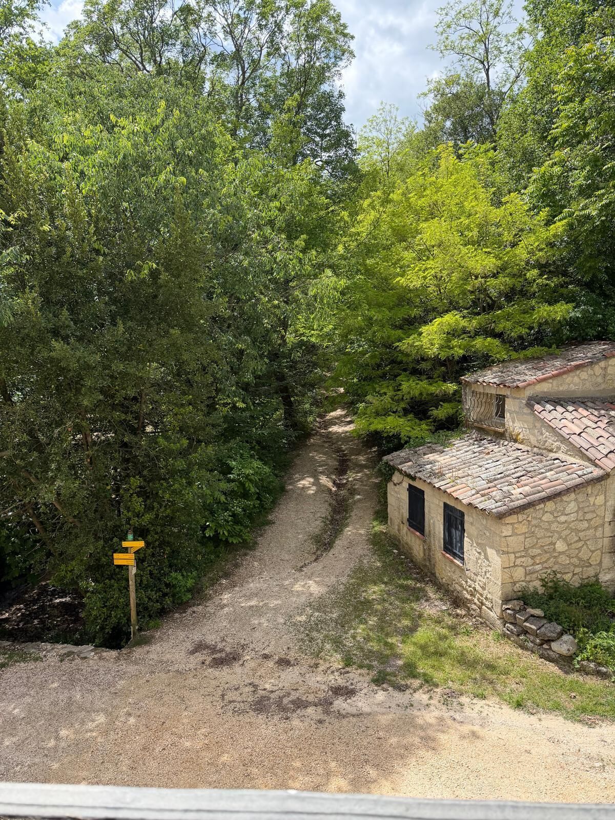 A rural path leading to a stone building with a tiled roof, surrounded by dense greenery.