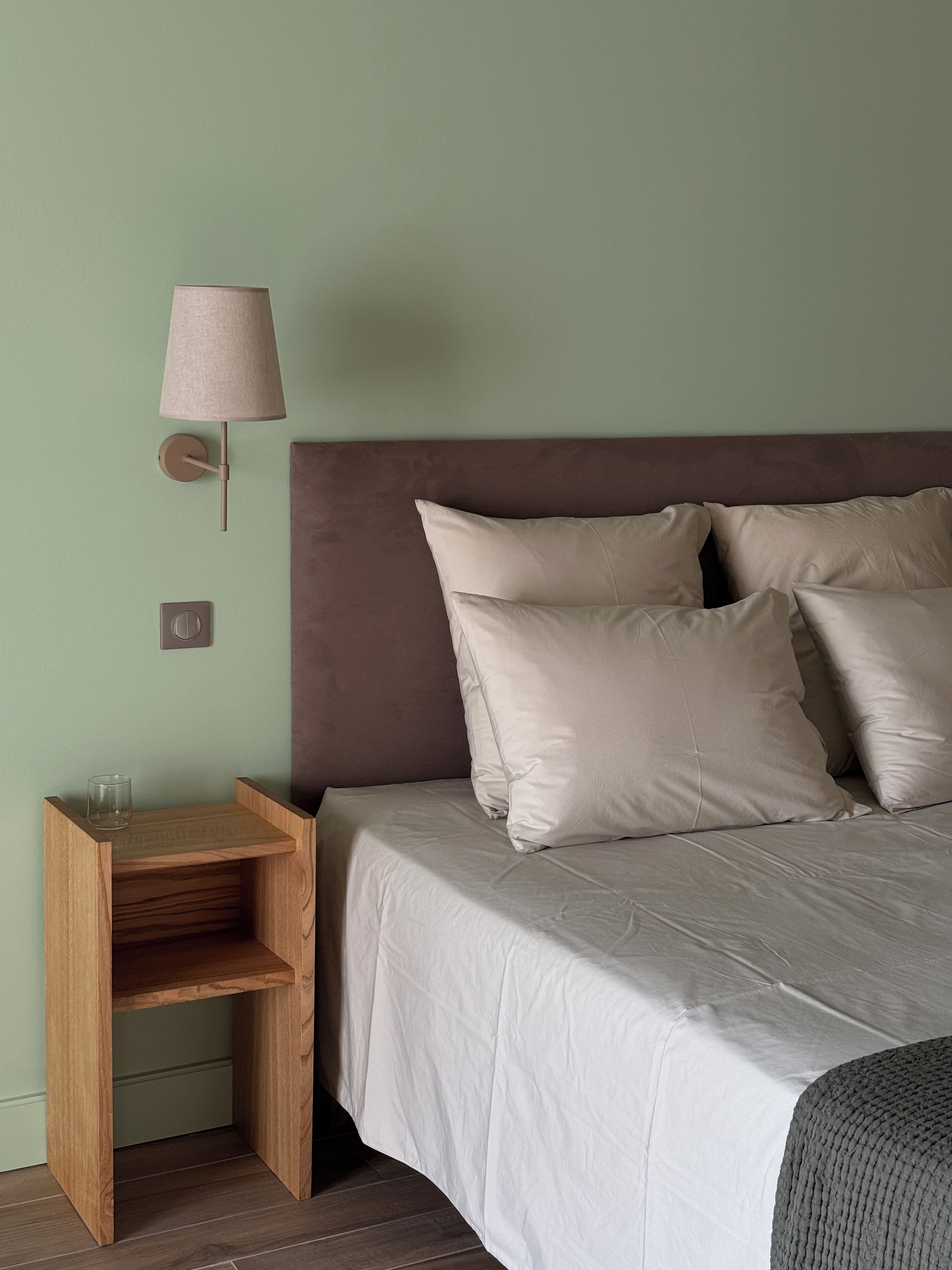 Uma cama com cabeceira, uma mesa de cabeceira com abajur e um interruptor fixado na parede.