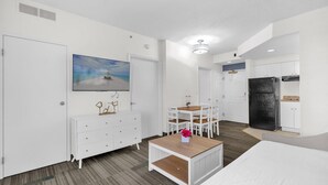 Ein modernes Hotelzimmer mit einem Bett, einem kleinen Tisch mit einer Vase, einem Bild von einer Strandansicht, einer Küchenzeile und einem Badezimmer.