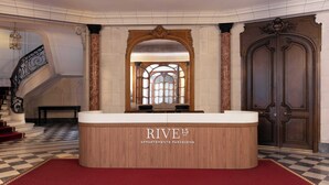 Vastaanottotiski, jossa on kyltti, jossa lukee ”RIVE 15 APARTMENTS PARISIENS”, ruutukuviollinen lattia, portaikko ja suuri puuovi.