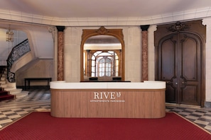 Ein Empfangstresen mit der Beschilderung 'RIVE 15 APARTMENTS PARISIENS', ein Schachbrettmuster-Boden, eine Treppe und eine große Holztür.