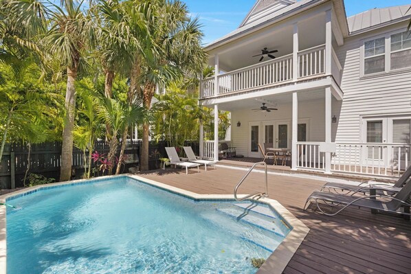A swimming pool with a wooden deck, lounge chairs, and a two-story house with a balcony. - Private Paradise: 3-bedroom Home with Pool and Tropical Backdrop (Duck Key)