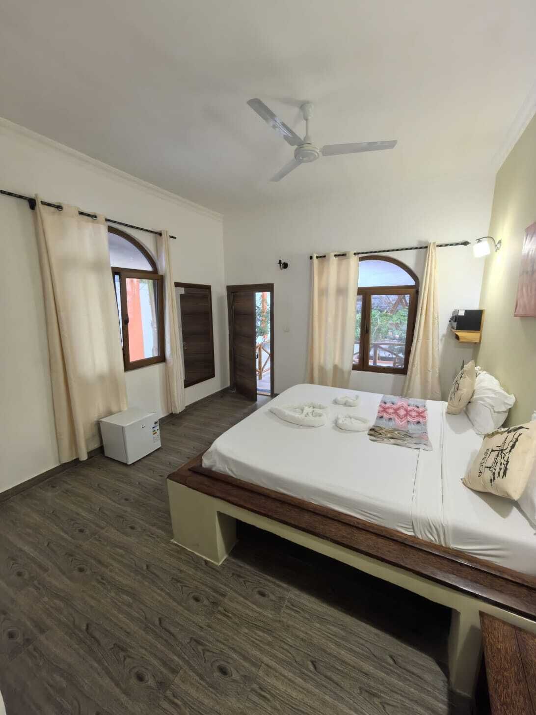 Un dormitorio con cama, ventilador de techo, ventana con cortinas y una mesita de noche.