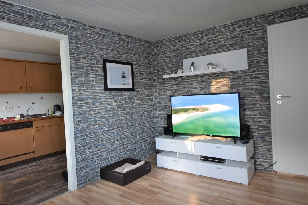 A modern living room with a stone-patterned wall, a flat-screen TV, a white entertainment unit, and a kitchen visible through an open doorway.