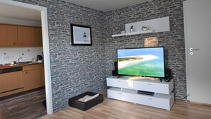 Ein modernes Wohnzimmer mit Wand im Steinoptik, einem Flachbildfernseher, einer weißen Unterhaltungsecke und einer durch eine offene Tür einsehbaren Küche.