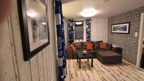 Ein Wohnzimmer mit einem braunen Ecksofa, einem kleinen Tisch, gemusterten Vorhängen, einem gerahmten Bild an der Wand und einer Wand aus Naturstein.