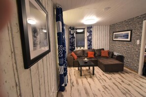 A living room with a brown sectional sofa, a small table, patterned curtains, a framed picture on the wall, and a stone accent wall. - NEW! Villa Totti - Lucas Stube (Neßmersiel)
