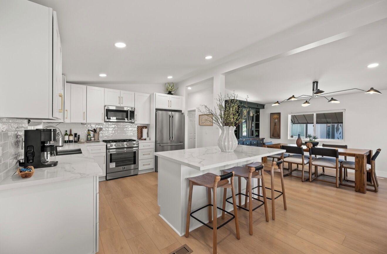 A modern kitchen with white cabinetry, stainless steel appliances, a marble island, and a dining area with wooden chairs.
