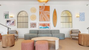 Een moderne woonkamer met een grijze bank, ronde poufs, een houten salontafel en wanddecoratie, waaronder een surfboard en abstracte schilderijen.
