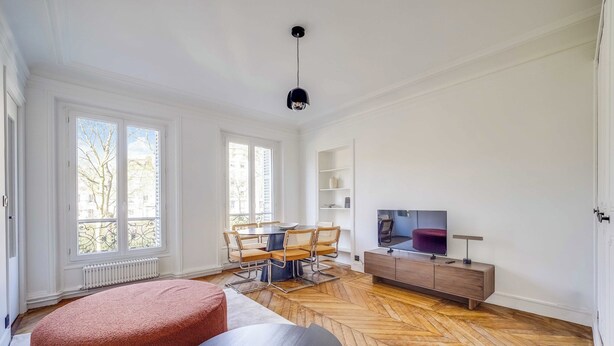 Ein geräumiger Wohnbereich mit Fischgrätparkett, einem Esstisch mit Stühlen, einer Couch, einem Fernseher und einer Holzkommode.
