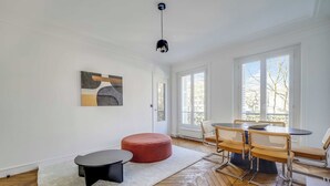 Ein modernes Wohnzimmer mit Fischgrätparkett, einem runden, roten Hocker, einem schwarzen Beistelltisch, einer Essecke mit rundem Tisch und Metallstühlen sowie einer schwarzen Pendelleuchte.