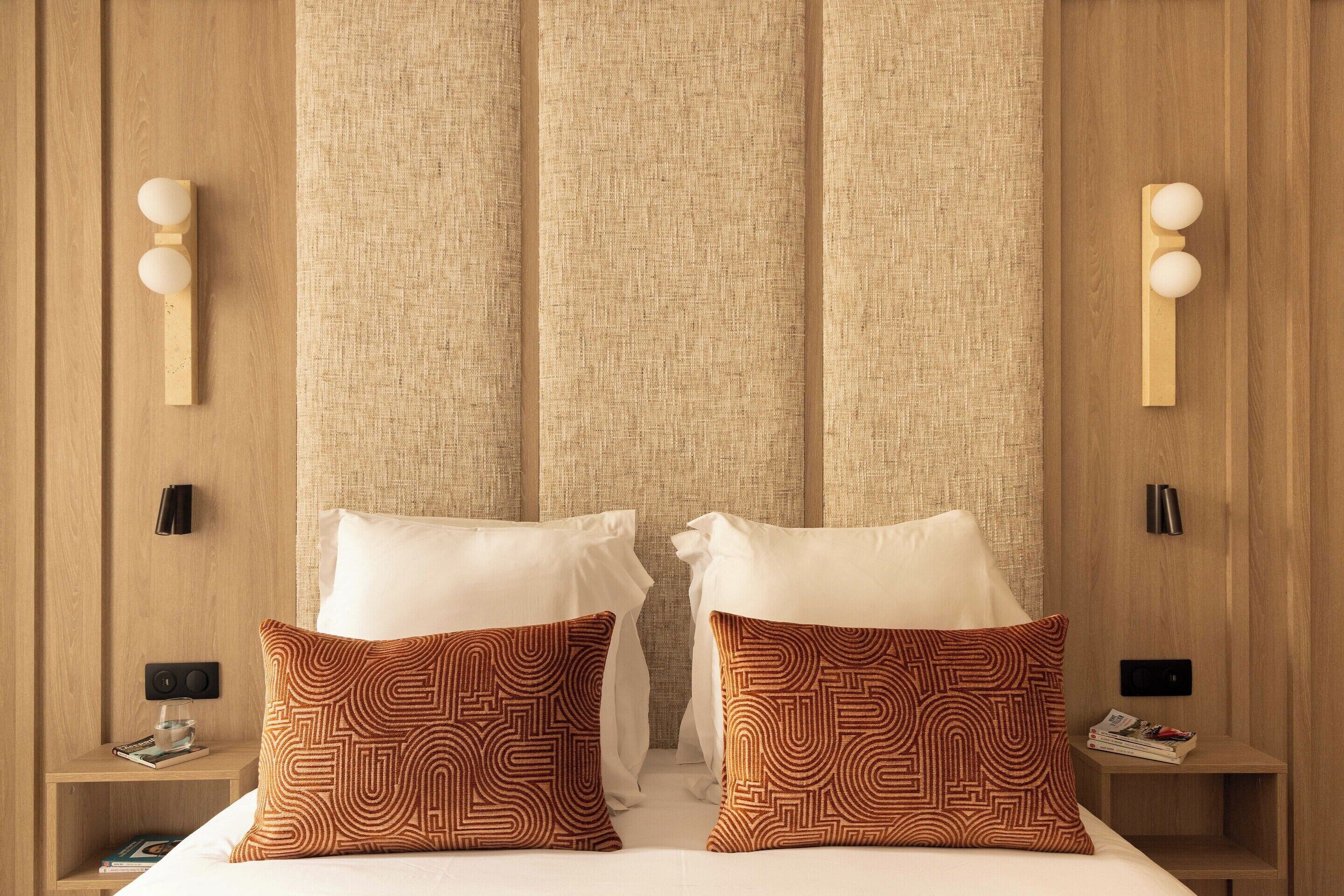 A hotel room with a bed, two patterned pillows, a nightstand with a lamp, and a wall-mounted light fixture.