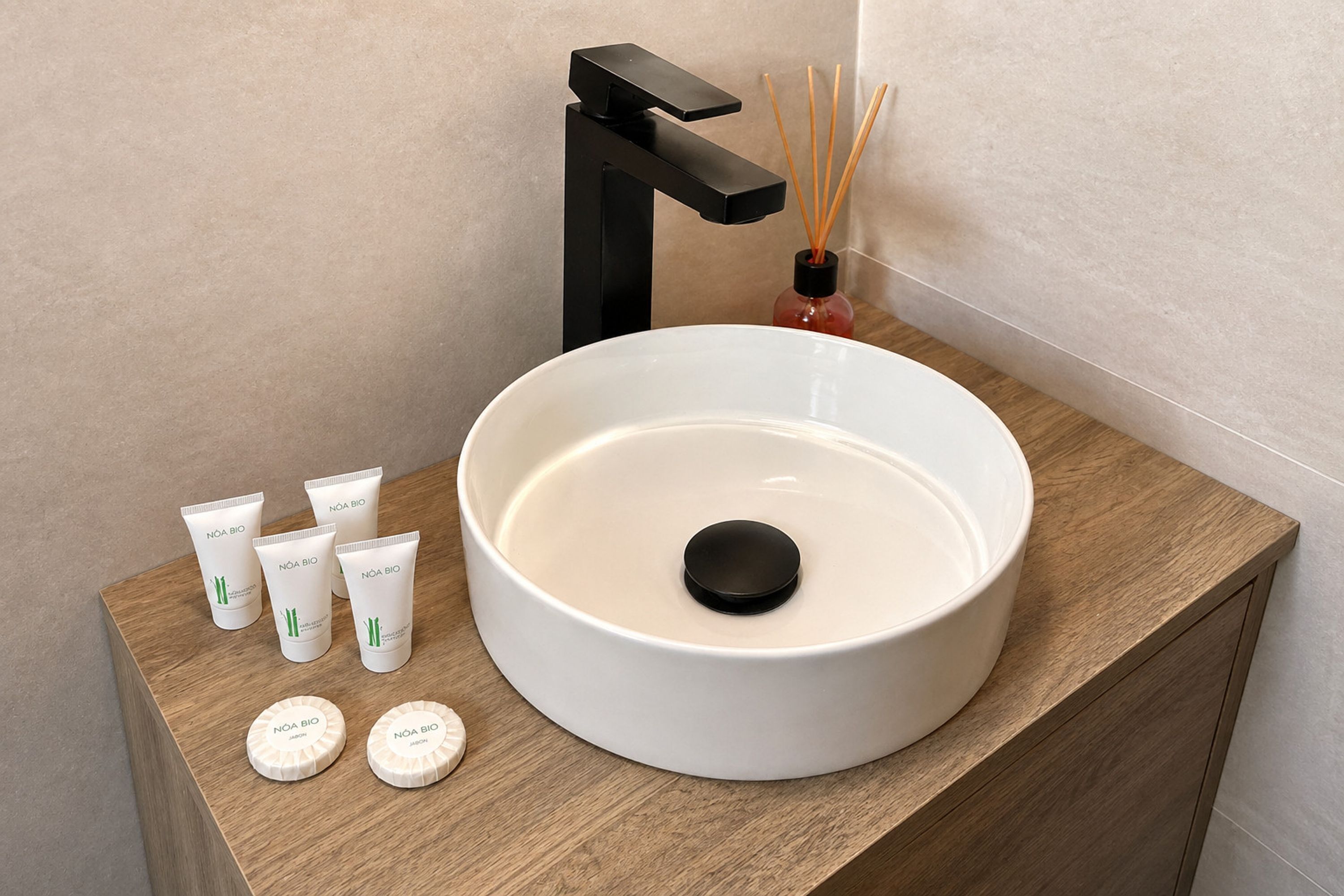 Een badkamer met een ronde witte wasbak, een zwarte kraan en een houten opbergmeubel. Op het opbergmeubel staan vijf Nôa Bio huidverzorgingsproducten.
