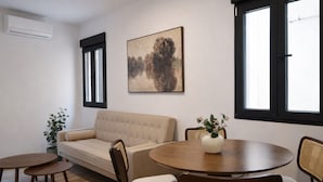 Una sala de estar con un sofá, una mesa de comedor redonda, sillas, una planta en maceta y un cuadro en la pared.