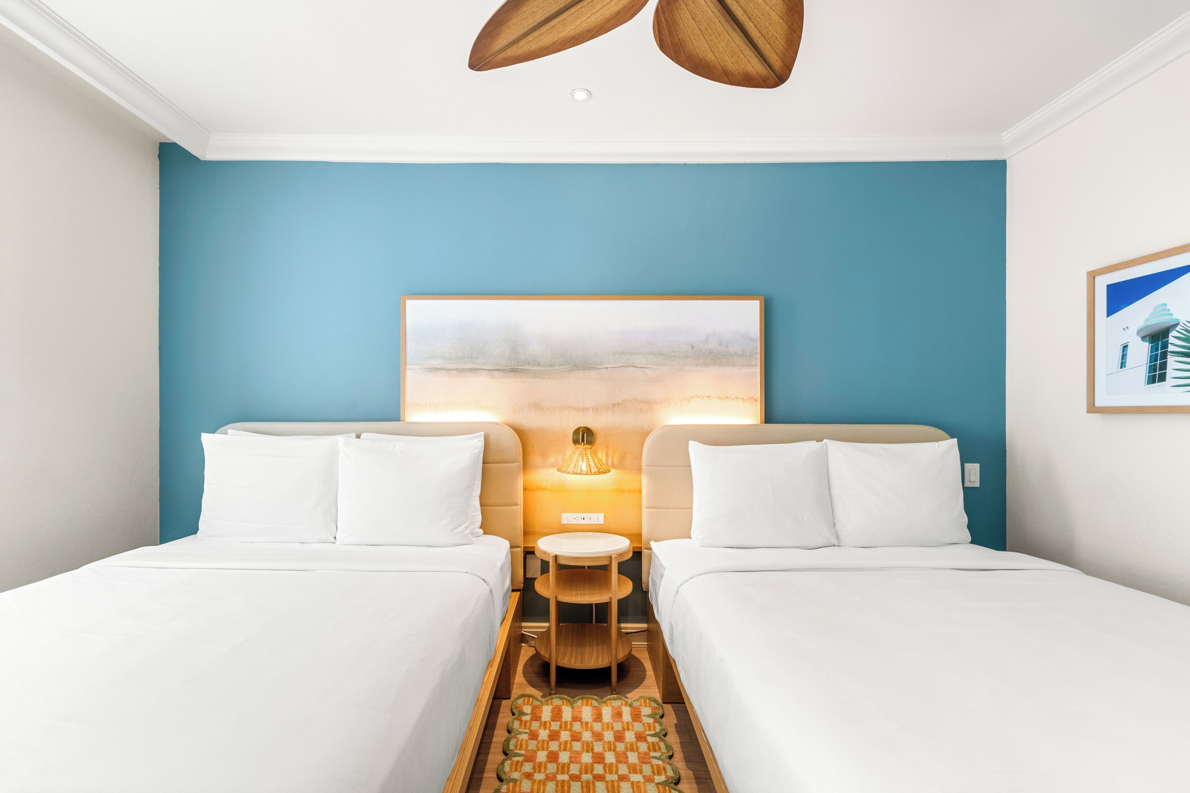 A hotel room with two beds, a small table, a lamp, and a painting on the wall.