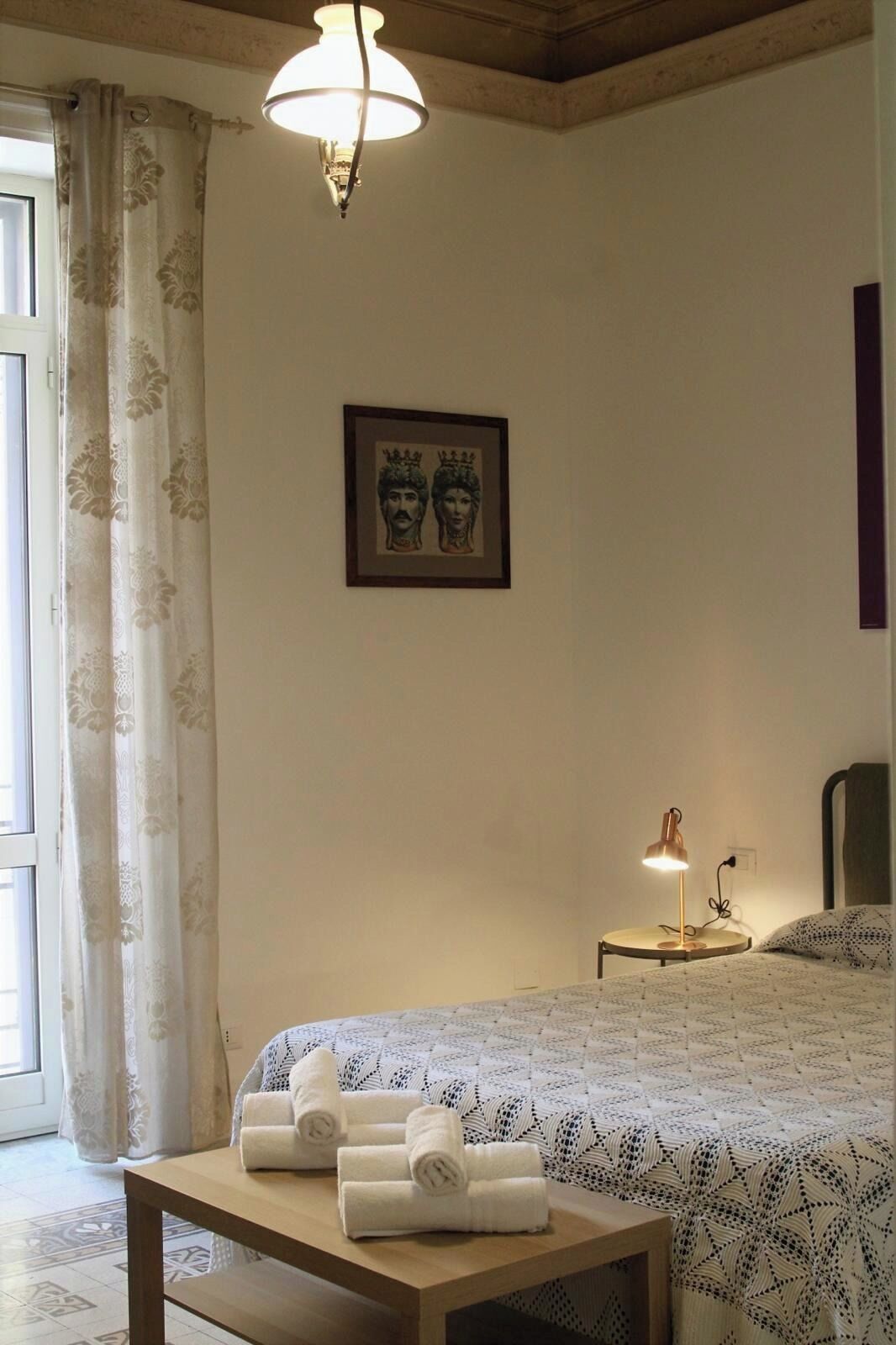 A bedroom with a bed, a table with towels, a nightstand with a lamp, a framed picture on the wall, and a window with curtains.