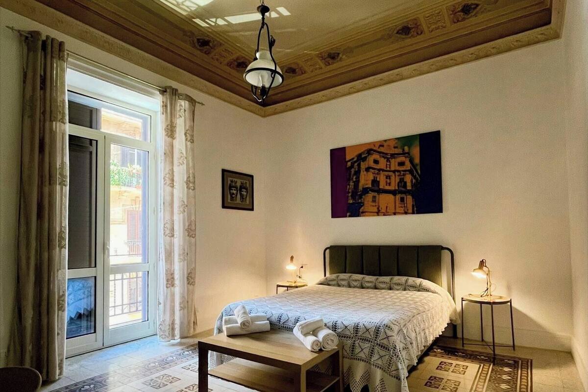 A bedroom with a bed, a nightstand, a hanging light fixture, a painting, and a patterned floor.