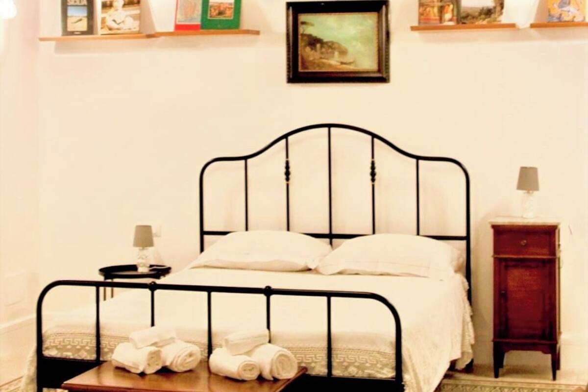 A bedroom with a bed, a nightstand, a shelf with books and pictures, and a chandelier.