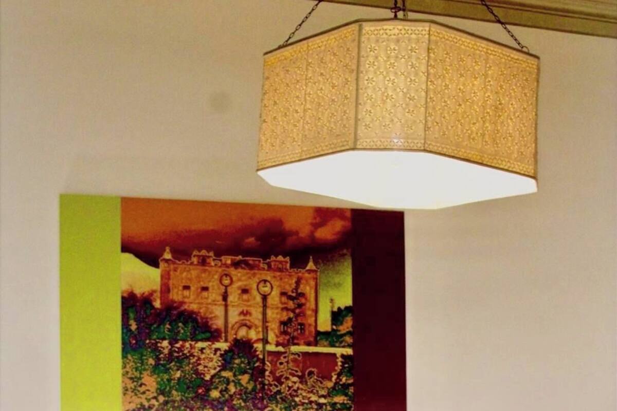 A bedroom with a bed, a hanging lamp, a wall-mounted painting, and a bedside table with a lamp.