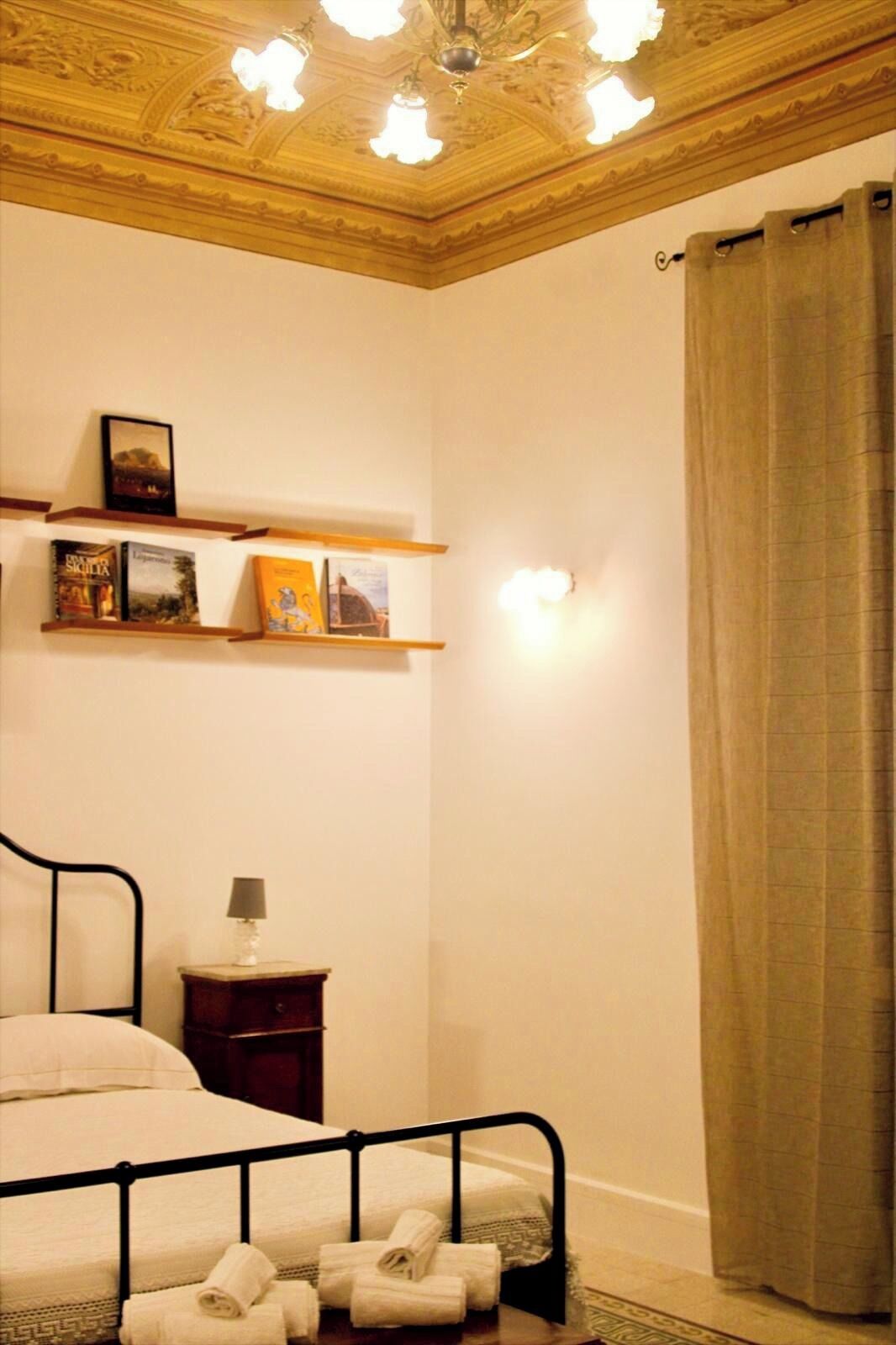 A bedroom with a bed, a nightstand, a lamp, a shelf with books, and a curtain.