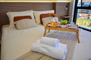 En snyggt bäddad säng med ett brickbord fullt av frukosttillbehör, inklusive croissanter, frukt och en kopp, i ett modernt hotellrum med utsikt.