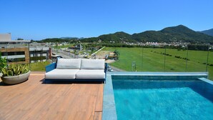 Un área de piscina en la azotea con un sofá blanco y una jardinera, con vistas a un paisaje urbano y montañas.