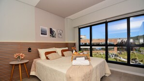 Un dormitorio moderno con una cama grande, una mesita de noche con un florero y vistas a un edificio a través de amplios ventanales.