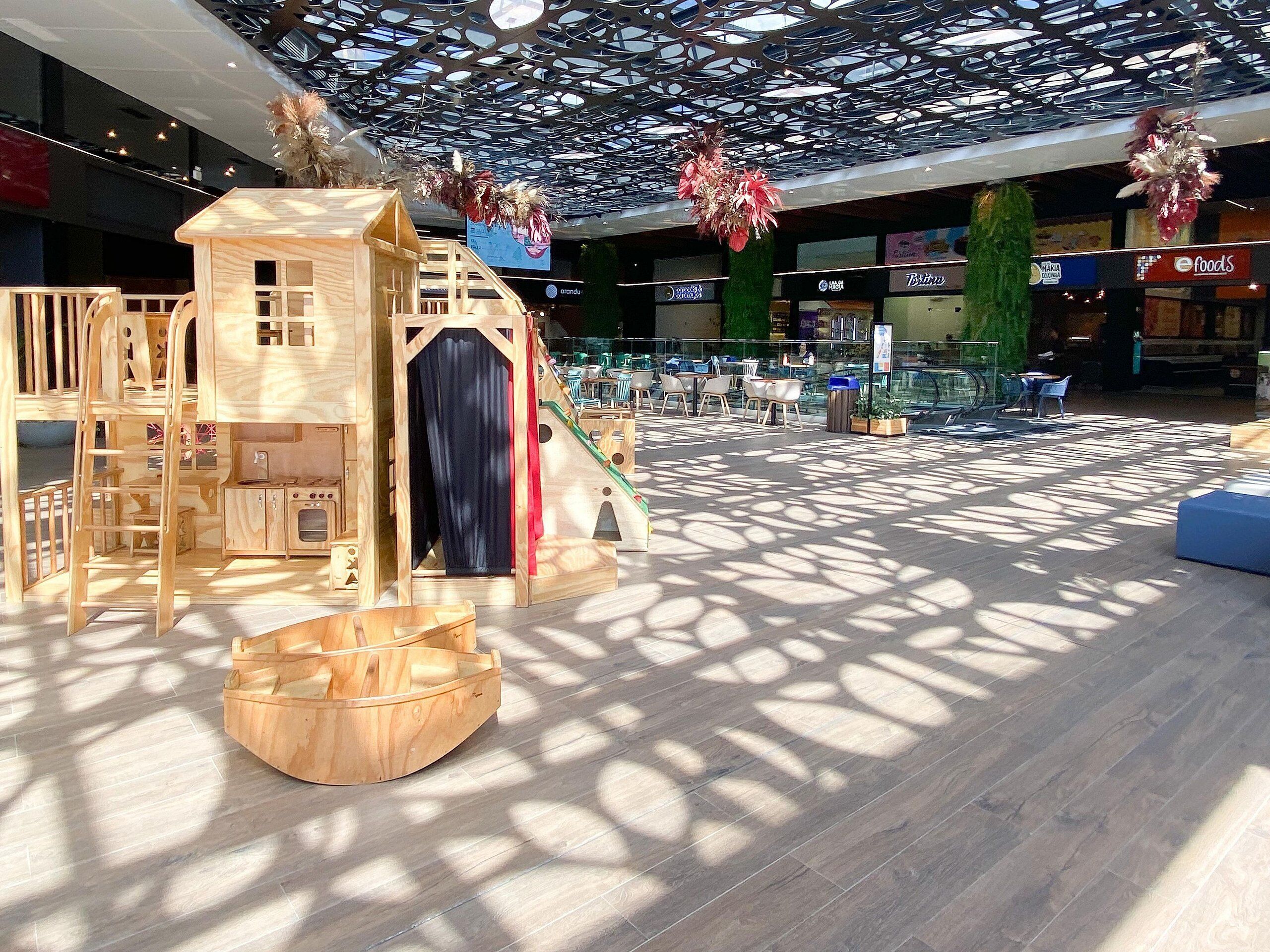 Zona de juegos infantiles con una casita de madera y tobogán, rodeada de mesas y sillas.