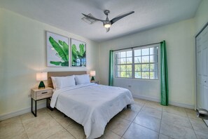 Un dormitorio con cama, mesitas de noche, ventilador de techo, ventana con cortinas y puerta.