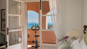 Una porta aperta che dà su un balcone con vista sul mare, una cyclette ferma e un letto con una lampada.