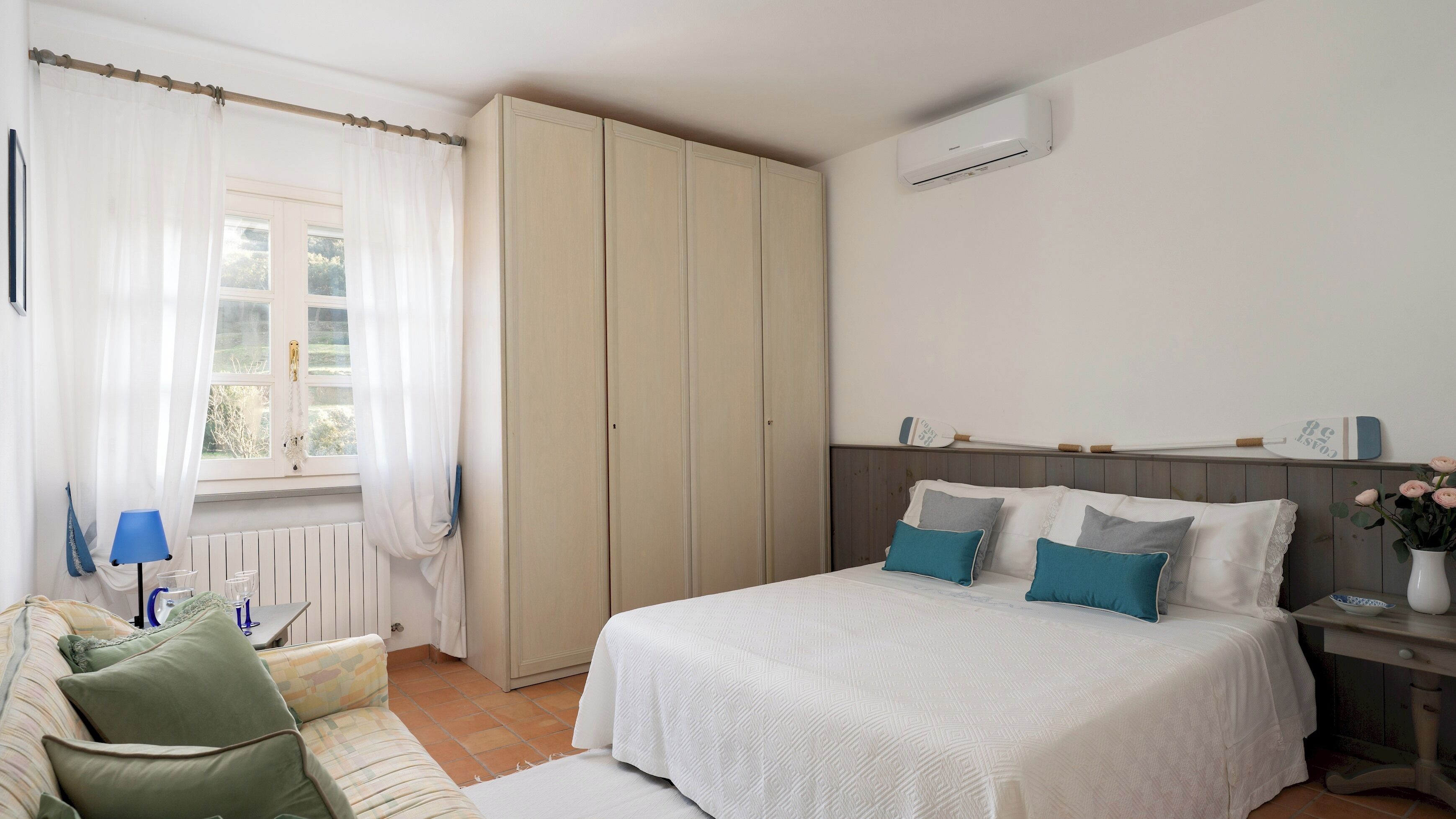 Une chambre à coucher comprenant un lit, une armoire, une fenêtre avec des rideaux, une table de chevet avec un vase et un climatiseur fixé au mur.