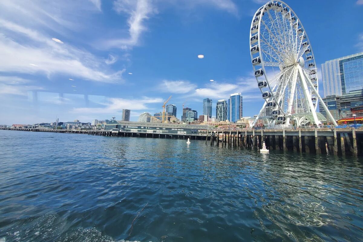 Un paisaje urbano frente al agua con una noria destacada, edificios modernos y un muelle que se extiende hacia el agua.