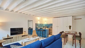 Ett modernt vardagsrum med en blå soffa, en platt-TV och en köksdel med en blå barstol.