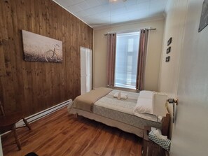 A bedroom with wooden walls, a bed, a window with blinds, a radiator, and a painting on the wall. - Auberge du vieux pont (Saint-Raymond)