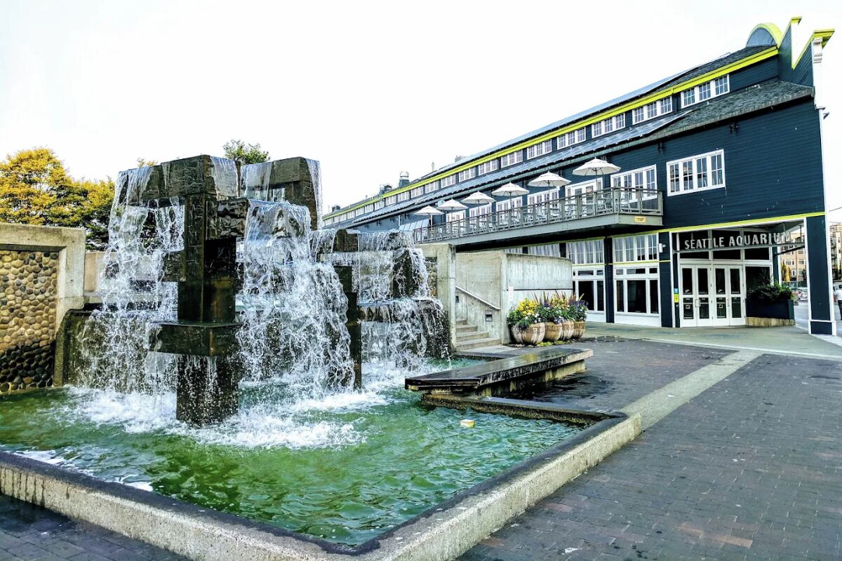 A public fountain with multiple tiers and water cascading down, a building labeled 'Seattle Aquarium' with a balcony, and a paved walkway.