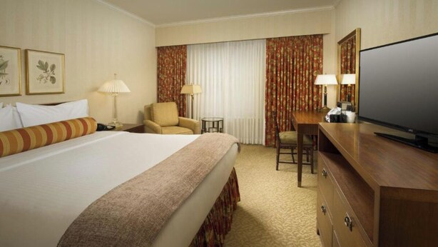 Une chambre d’hôtel avec un grand lit, deux fauteuils, un bureau avec une télévision et une grande fenêtre avec des rideaux.