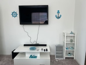 A room with a TV mounted on the wall, a white entertainment unit with a remote control, and a small white shelf with decorative items.