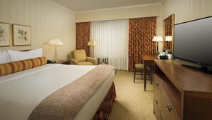 Ein Hotelzimmer mit einem großen Bett, zwei Sesseln, einem Schreibtisch mit Fernseher und einem großen Fenster mit Vorhängen.