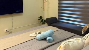 Una habitación de hotel con cama, televisor montado en la pared, ventana con persianas y una pequeña planta en un estante.