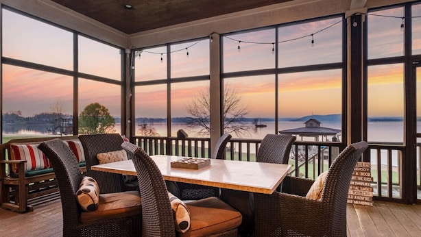 A covered patio with a table and chairs, large windows with a view of a lake and sunset, and a sign with rules. - Family Retreat ~ Waterfront Luxury Home Sleeps 26 Comfortably (Centre)