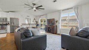 A modern living room with a grey sofa, a ceiling fan, a flat-screen TV, and a kitchen area with white cabinets and appliances. - Lakefront Getaway w Boat Slip + Patio - Sleeps 8 - Fully Stocked - EV charger (Hot Springs)