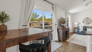 A modern living room with a wooden dining table, a black upholstered chair, a blue sofa, a small side table, a mirror, and a view of a dock and boats through large windows. - Lakefront Getaway w Boat Slip + Patio - Sleeps 8 - Fully Stocked - EV charger (Hot Springs)