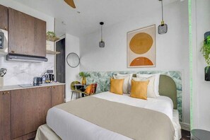 A modern hotel room with a bed, a desk, a kitchen area, and a wall-mounted artwork. - Charming Studio Apartment - AC- 2P - Nation (Paris)