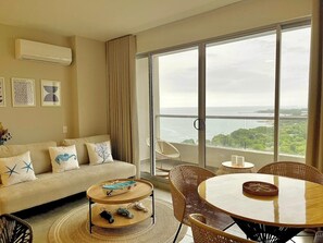 A living room with a sofa, coffee table, dining table, and chairs, with a large window offering a view of the sea. - Luxury Apartment in Santa Marta | Ocean View (Santa Marta)