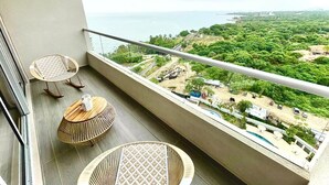 Een balkon met twee rieten stoelen en een tafel, met uitzicht op een kustlandschap met begroeiing en gebouwen.